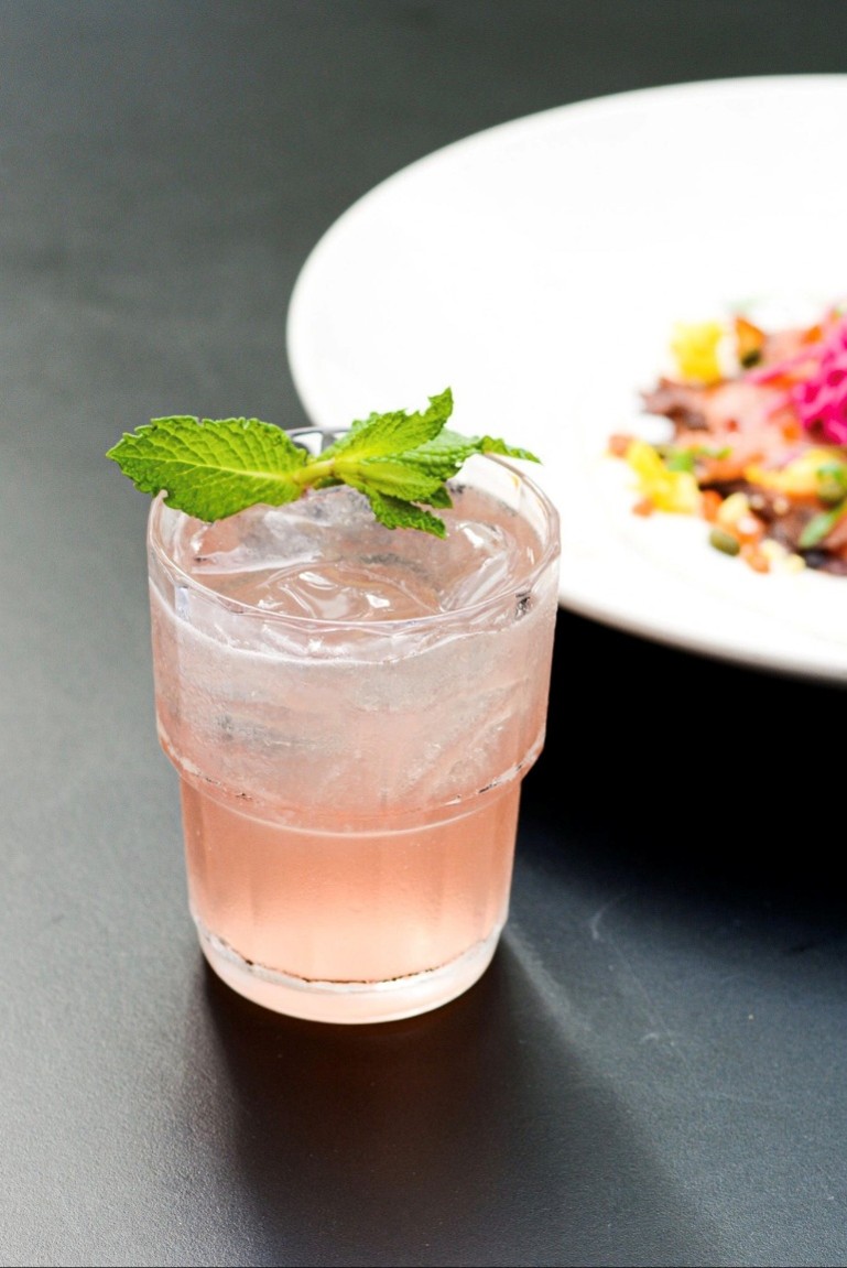 Pink craft cocktail garnished with fresh mint beside a plated dish at Blueprint on 3rd in Birmingham